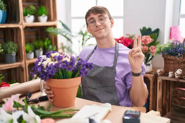 花屋で働く白人の金髪の男は 勝利のサインをしている指を見ているカメラに向かって微笑んでいます ナンバー2 — ストック写真