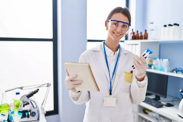 Young caucasian woman scientist holding test tubes reading notebook at laboratory