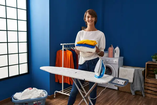 Brunette woman holding folded laundry after ironing smiling with a happy and cool smile on face. showing teeth.