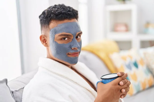Young latin man relaxed on sofa with facial mask treatment drinking coffee at home