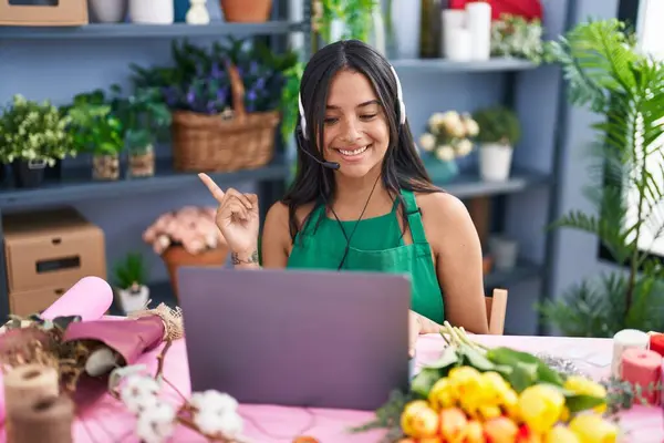 Brunette woman working at florist shop doing video call smiling happy pointing with hand and finger to the side