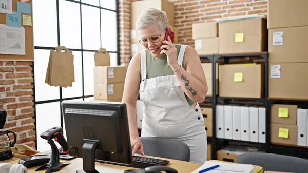 中年の灰色の髪の女性Eコマースビジネスワーカーは オフィスで働くコンピュータを使用してスマートフォンで話します — ストック写真
