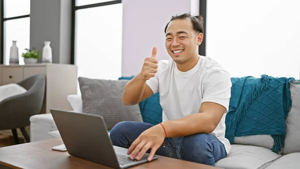 Cheerful young chinese guy happily sitting on home sofa, giving the thumbs up gesture while using a laptop, immersed in the joy of the internet technology within the comfort of his living room