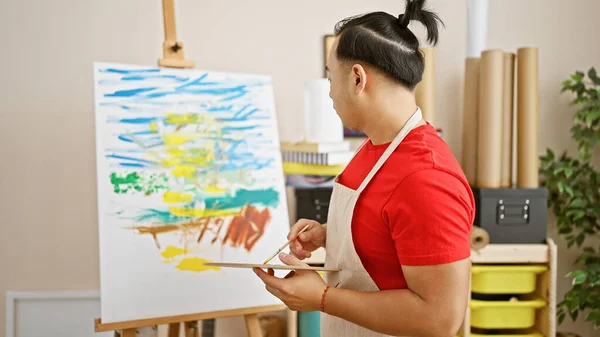 Handsome young chinese man, confident artist, drawing indoors, immersed in creativity at art studio with paintbrush in hand, adorned with traditional pigtail hairstyle & apron