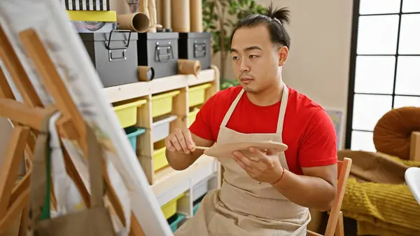 Serious young chinese man artist, relaxed yet concentrated, draws at his studio. handsome with pigtail hairstyle and apron, mastering the art on canvas with paintbrushes, embracing creativity.