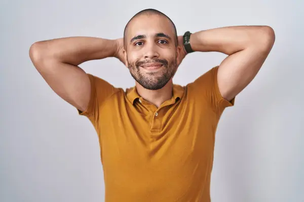 Hispanic man with beard standing over white background relaxing and stretching, arms and hands behind head and neck smiling happy