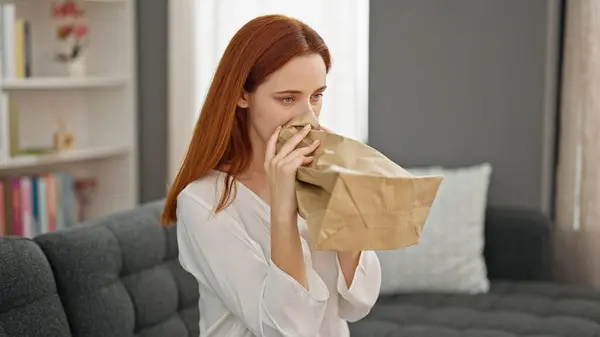 Young redhead woman stressed sitting on sofa blowing air on paper bag at home