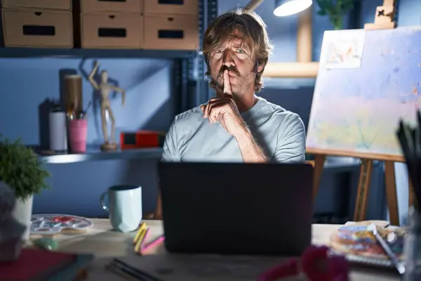 中年男子坐在艺术工作室里 晚上拿着笔记本电脑 要求安静 手指放在嘴唇上 沉默和秘密概念 — 图库照片