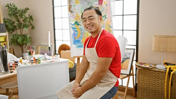 Cheerful young chinese art student, a gifted painter, radiates confidence sitting on his chair, smiling in the middle of his art academy\'s indoor studio, brushes and paint palette in hand