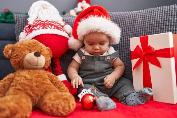自宅でクリスマスの帽子をかぶっているソファーに座っている愛らしいヒスパニックの幼児 — ストック写真