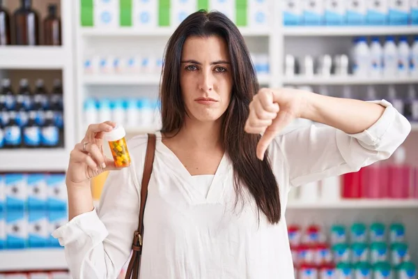 Young brunette woman shopping at pharmacy drugstore holding pills with angry face, negative sign showing dislike with thumbs down, rejection concept