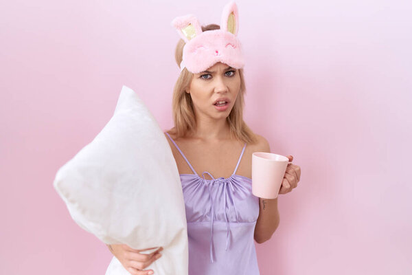 Young caucasian woman wearing pajama hugging pillow and drinking coffee in shock face, looking skeptical and sarcastic, surprised with open mouth 