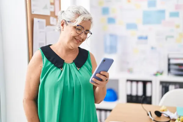 Middle age grey-haired woman business worker smiling confident using smartphone at office