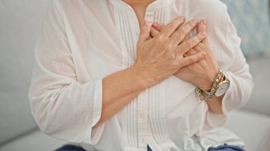 Middle age woman sitting on sofa with hands on heart at home