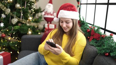 Evdeki Noel ağacının yanında akıllı telefon kullanan genç sarışın kadın.