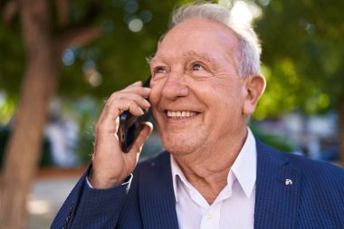 Orta yaşlı, kır saçlı, gülümseyen, kendine güvenen, parkta akıllı telefondan konuşan bir adam.