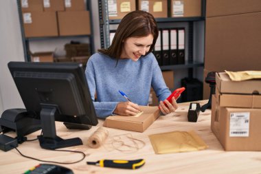 Genç sarışın kadın ecommerce iş işçisi. Ofiste paket üzerinde akıllı telefon yazıları kullanıyor.