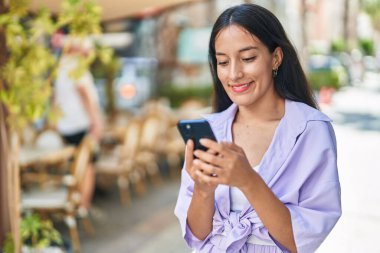 Genç, güzel, İspanyol bir kadın sokakta akıllı telefon kullanarak gülümsüyor.