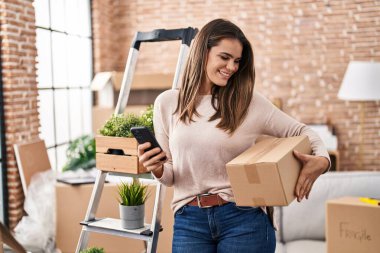 Young beautiful hispanic woman using smartphone holding package at new home