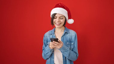 Kafkasyalı genç bir kadın noel şapkası takıyor ve izole edilmiş kırmızı arka plan üzerinde akıllı telefon kullanıyor.