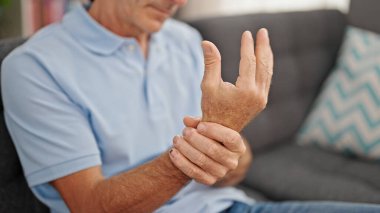 Orta yaşlı bir adam koltukta oturuyor ve evinde bilek ağrısı çekiyor.