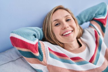 Sarışın genç kadın ellerini başının üstüne koyup evdeki kanepede oturuyor.