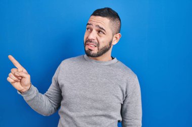 Mavi arka planda dikilen İspanyol adam endişeli ve endişeli bir ifadeye işaret ediyor. 