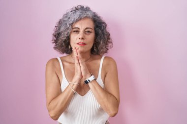 Middle age woman with grey hair standing over pink background begging and praying with hands together with hope expression on face very emotional and worried. begging. 