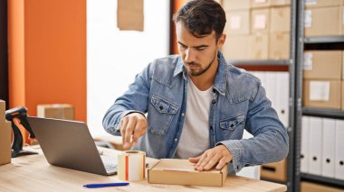 Young hispanic man ecommerce business worker packing cardboard box at office