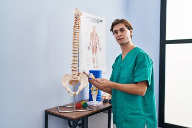 Young caucasian man physiotherapist touching anatomical model of spinal column at rehab clinic