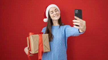 Kafkasyalı genç bir kadın noel şapkası takıyor ve izole edilmiş kırmızı arka planda akıllı telefon hediyesiyle selfie çekiyor