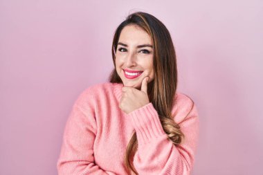 Pembe arka planda duran genç İspanyol kadın kameraya kendinden emin bir şekilde bakıyor kollarını kavuşturmuş ve çenesini kaldırmış gülümsüyor. Olumlu düşünmek. 