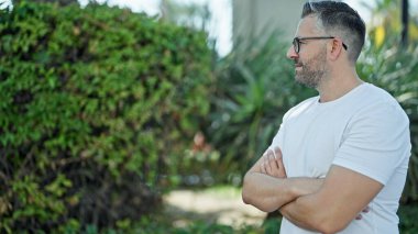 Parkta yüzü ve kolları çaprazlanmış beyaz saçlı bir adam duruyor.