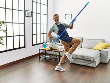 Middle age grey-haired man using mop as a guitar at home