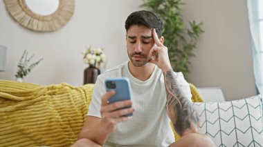 Genç İspanyol adam akıllı telefon kullanıyor. Evde üzgün bir ifadeyle kanepede oturuyor.