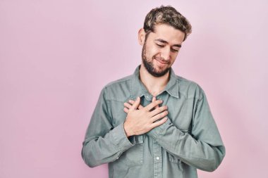 Pembe arka planda duran sakallı, elleri göğsünde, gözleri kapalı ve yüzünde minnet dolu bir jest olan İspanyol bir adam. sağlık kavramı. 