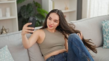 Evdeki kanepede oturan ve akıllı telefonuyla selfie çeken genç, güzel İspanyol kadın.