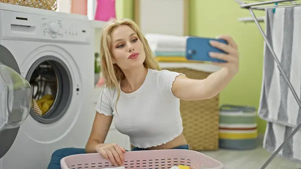 若い金髪の女性は洗濯室でスマートフォンでセルフィーを作ります — ストック写真