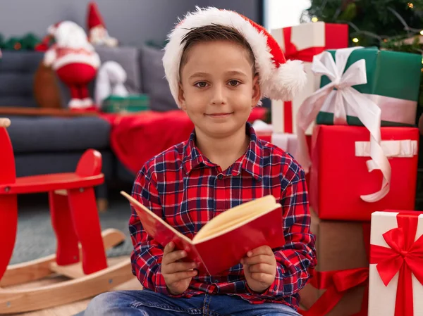 自宅でクリスマスツリーで床に座って本を読む愛らしいヒスパニックの少年 — ストック写真