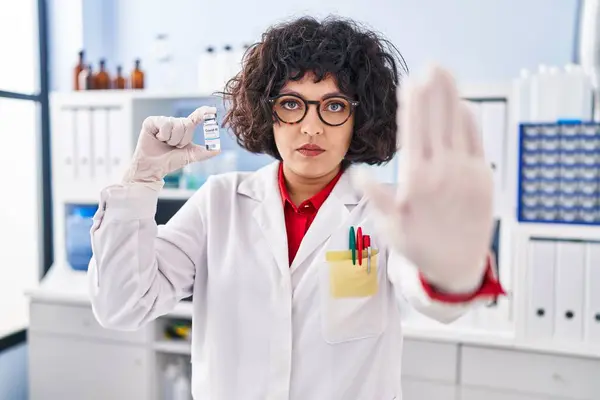 オープンハンドでワクチンを保持するカーリーヘアを持つヒスパニックの医師女性は 深刻で自信を持った表現 防衛ジェスチャーで停止サインを行います — ストック写真