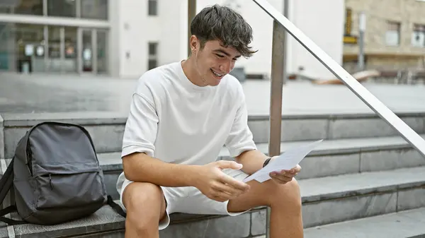 魅力的な学生であるヒスパニック系の若者を魅了し キャンパスの階段でリラックスしながら教育文書を読み 活発な都市の大学の設定で暖かい日光の下で自信を持って微笑んでいます — ストック写真