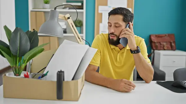 Young hispanic man being fired sitting on table talking on smartphone at the office