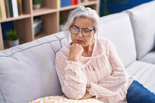 Middle age grey-haired woman sitting on sofa with serious expression at home