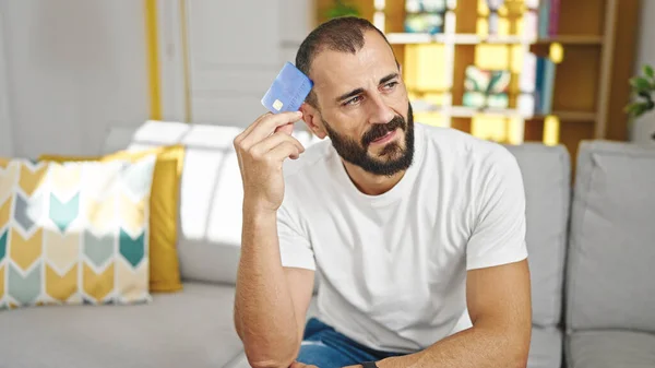 Jovem Hispânico Segurando Cartão Crédito Sentado Sofá Pensando Casa — Fotografia de Stock