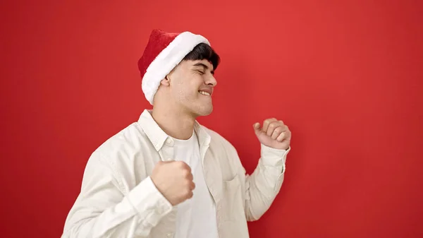 若いヒスパニックの男は 孤立した赤い背景の上に踊るクリスマスの帽子を着て自信を持って微笑みます — ストック写真
