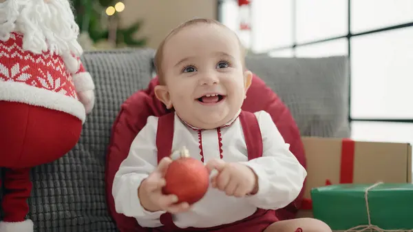愛らしいHispanic赤ちゃん保持クリスマス装飾ボール座っています上のソファにホーム — ストック写真