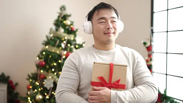Young chinese man listening to music holding gift by christmas tree at home