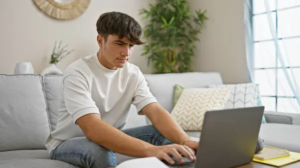 熱心な若いヒスパニック系男性学生は オンライン教育に魅了され 居心地の良いリビングルームのソファーに座って ノートパソコンの画面に焦点を当て テクノロジーに囲まれて自宅でリラックス — ストック写真