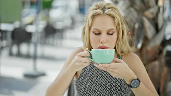 Young blonde woman drinking cup of coffee sitting on table at coffee shop terrace