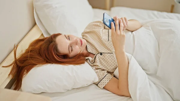 年轻的红头发女人用智能手机躺在卧室的床上 — 图库照片